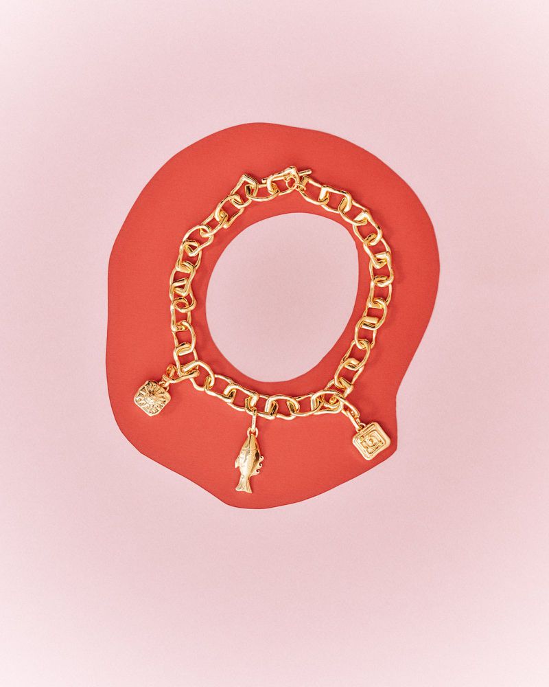 Gold charm bracelet featuring charms shaped like a heart, fish, and logo against a salmon background, jewelry concept, studio shot.