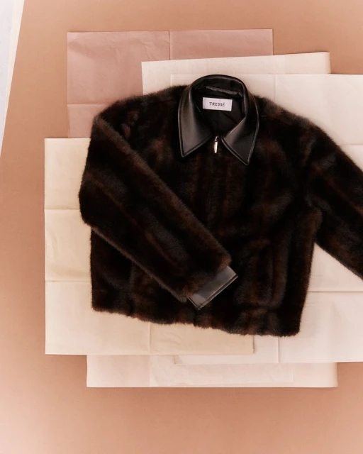 Dark brown fur jacket with a leather collar on top of folded white and pink paper layers, fashion, studio shot, product photography.