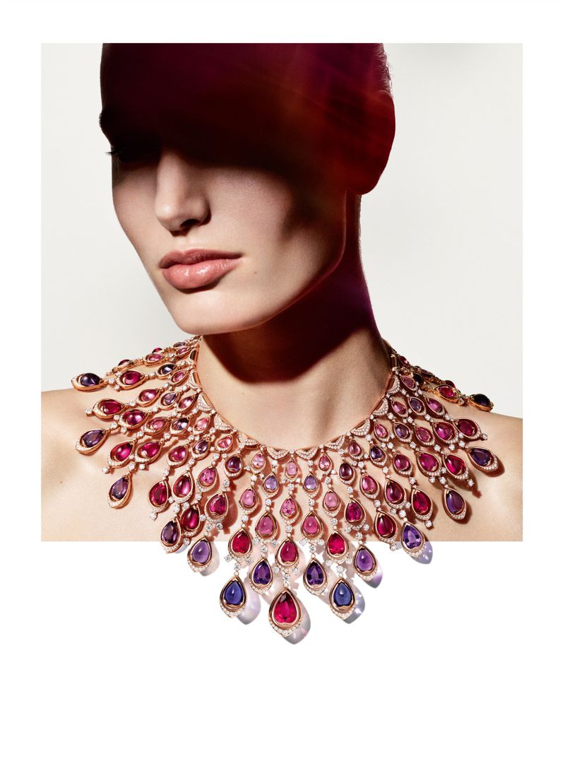 Woman in a beauty shot wearing an elaborate statement necklace with colorful gemstones against a bright, neutral background in a studio.