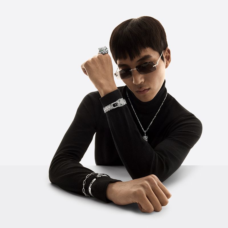 Man wearing jewelry poses indoors against a white backdrop. He's wearing sunglasses, a black turtleneck, a ring, bracelet, and necklace.