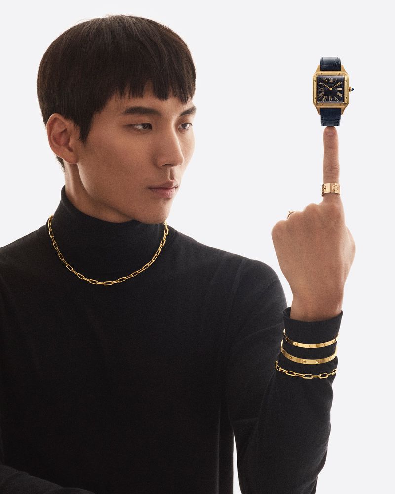 Elegant Asian man in black turtleneck with gold chain necklace, posing with a gold wristwatch on his fingertip against white backdrop.