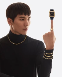 Man in black turtleneck balancing a luxury gold watch on his raised index finger against a white background