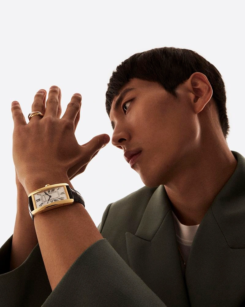 Man wearing a gold watch and ring with an olive green blazer against a plain white background