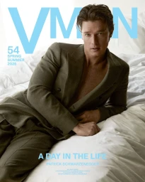 Patrick Schwarzenegger wearing an olive green suit jacket lying on white bedsheets