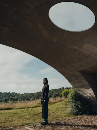 A woman in a black suit stands in a grassy field beneath a modern concrete arch with a circular opening to the sky above.