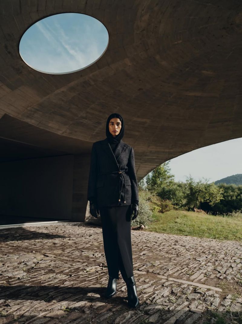 A woman wearing a dark jacket and skirt stands outdoors in front of a modern concrete structure on a stone surface, with a clear sky.