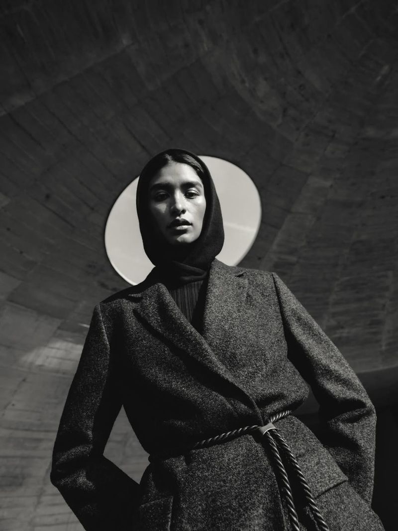 Monochrome portrait of a stylish adult female in a coat with a rope belt and headscarf, set against a concrete architectural background.