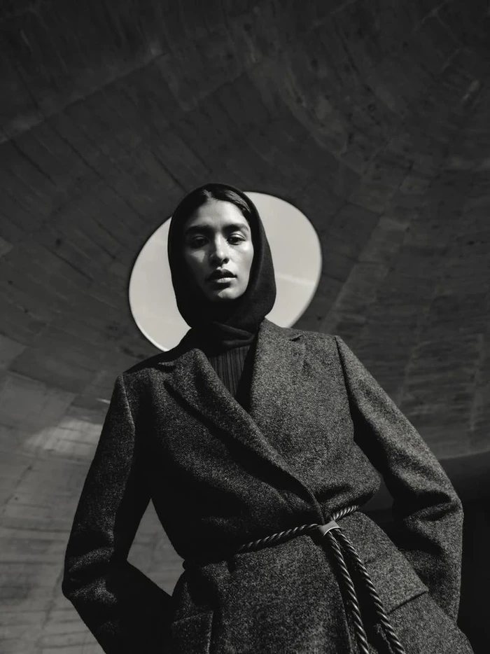 Model wearing a dark wool coat and hood looking down against a textured stone archway for Agnona Fall Winter 25.
