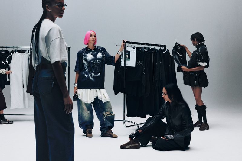 Five people in a light studio setting with clothing racks filled with dark apparel, modelling relaxed fashion looks.