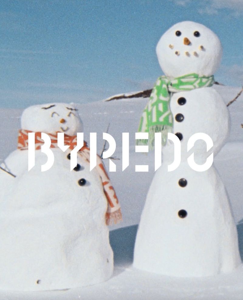 Two snowmen wearing scarves stand in a snowy landscape under a blue sky, with the BYREDO logo superimposed on the lower left.
