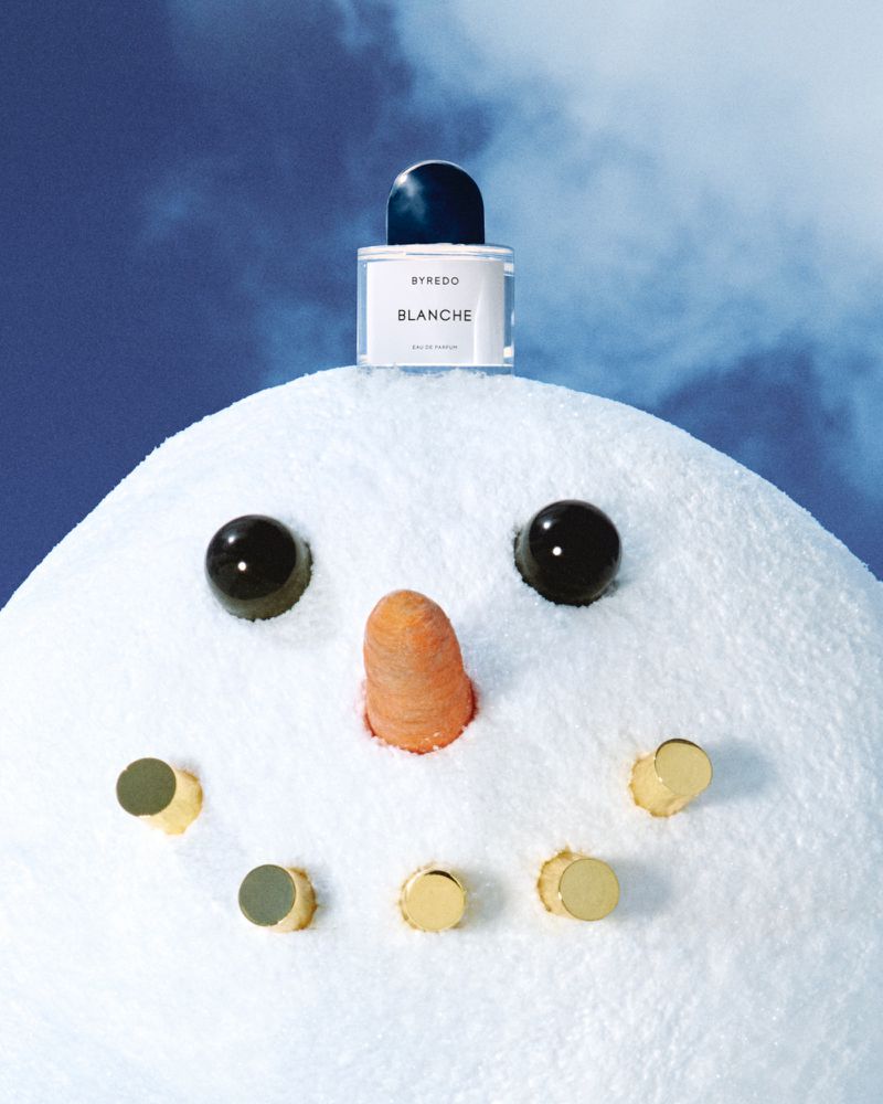 Byredo Blanche perfume bottle sits atop a plush snowman head with carrot nose and button eyes in a studio shot with a blue background.