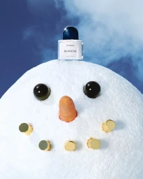 Byredo Blanche perfume bottle resting on a snowman head against a bright blue winter sky.