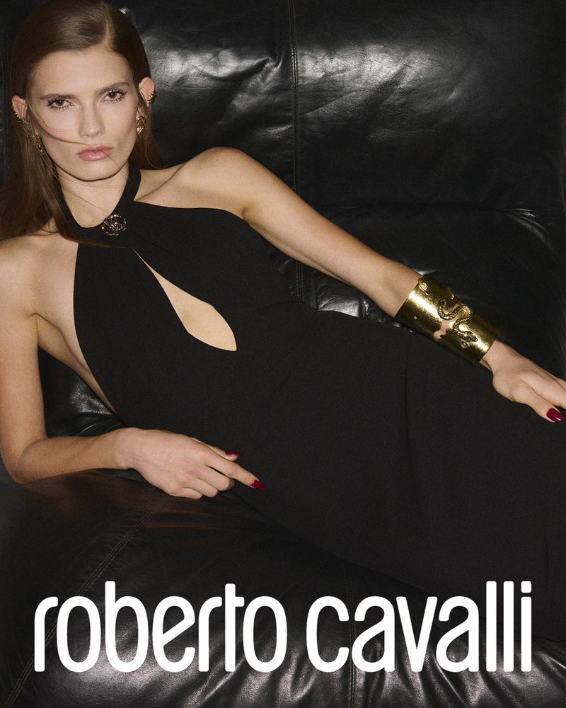 Glamorous adult female model in a black halter dress with a gold cuff bracelet poses on a black leather couch with soft, diffused lighting.