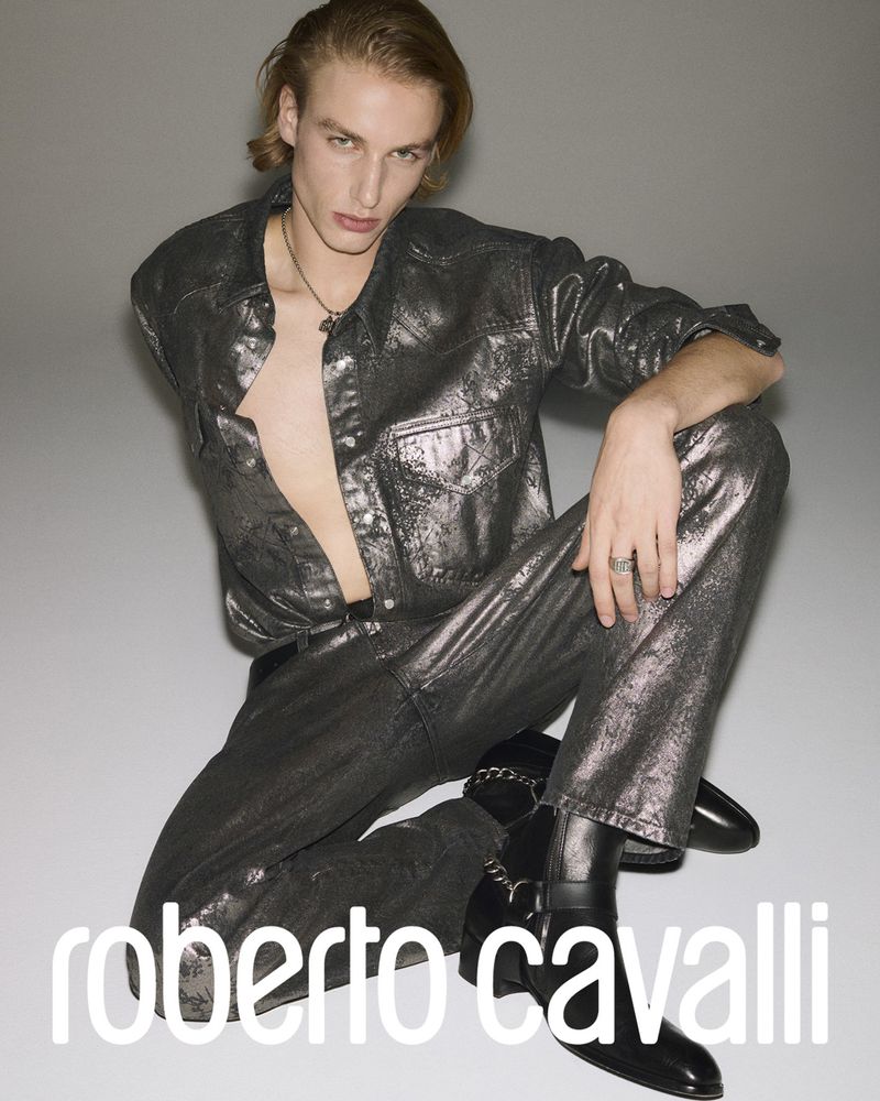 A young adult man poses indoors wearing a shiny silver outfit consisting of a shirt and pants with black boots on a gradient background.