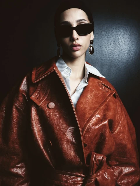 Portrait of a woman with a headscarf, sunglasses, and large earrings, wearing a red-brown leather coat against a dark, grainy background.