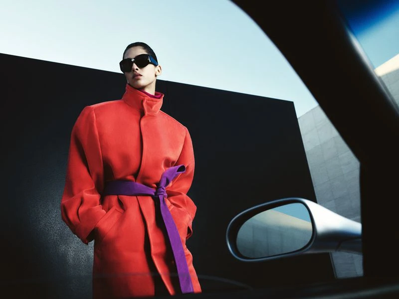 Stylish adult model in sunglasses poses in an orange coat tied with a purple sash near a modern building and car in daylight.