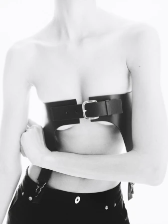 Close-up of a slender woman with a bare torso and black trousers wearing a black leather harness with a silver buckle, shot in black and white.