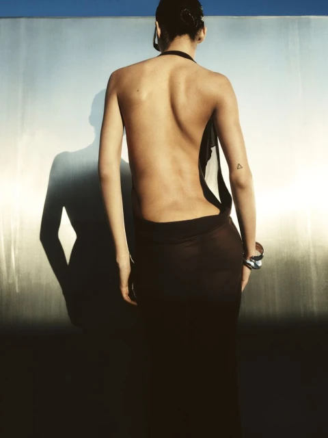 Back view of adult female fashion model in brown dress with open back, silver bracelet in photo studio with silver wall and blue sky.