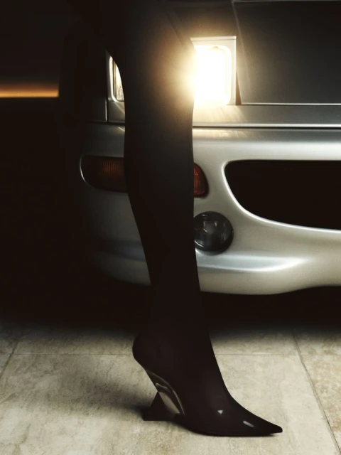 A leg in black hosiery and a geometric black heel stands in front of a silver car, highlighting fashion and automotive elements.