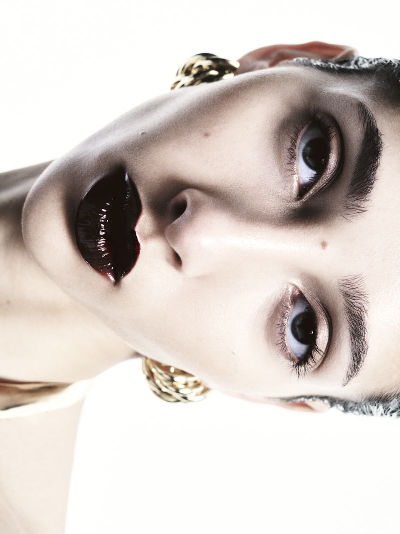 Close-up portrait of a woman with dramatic dark lipstick and statement earrings, against a stark white background in a studio setting.