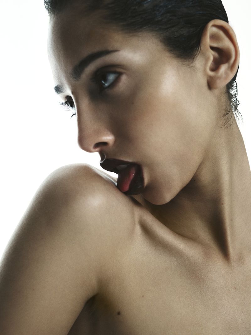 Close-up portrait of a bare-shouldered woman with dark lipstick licking her shoulder in a brightly lit, studio environment.