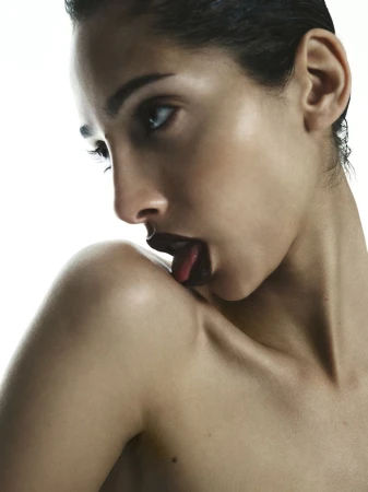 Close-up portrait of a bare-shouldered woman with dark lipstick licking her shoulder in a brightly lit, studio environment.