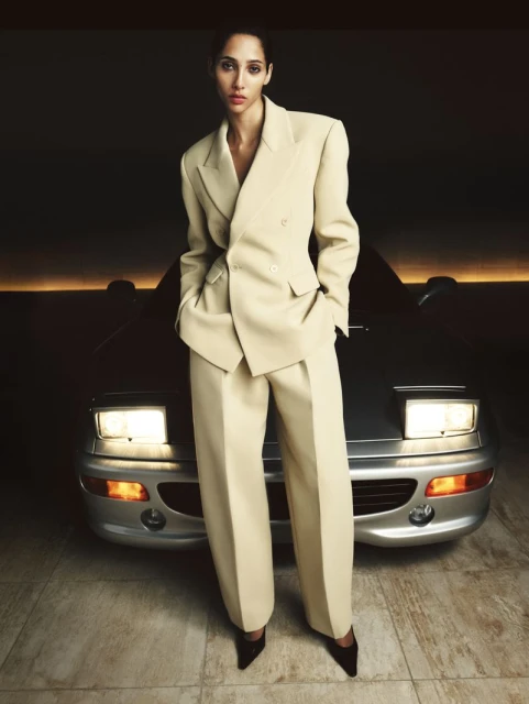 A woman in a beige suit poses in front of a silver sports car, with soft lighting enhancing the car's sleek design and her stylish attire.