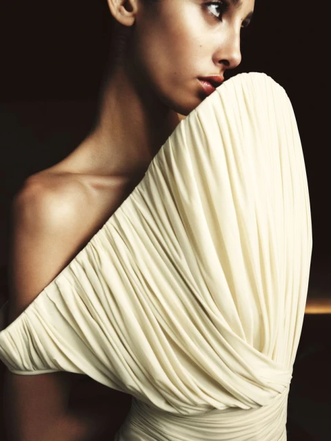 Glamorous portrait of a young woman in an off-the-shoulder ivory gown, looking off-camera against a dark, moody background.