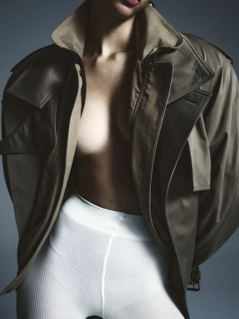 A fashion shot of a woman wearing a tan jacket with no top and white leggings, posing against a grey background.