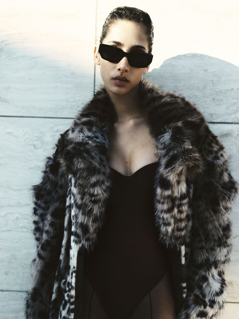 Stylish adult woman wearing a leopard print coat and sunglasses stands against a textured wall in a fashion forward portrait.