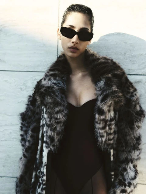 Stylish adult woman wearing a leopard print coat and sunglasses stands against a textured wall in a fashion forward portrait.