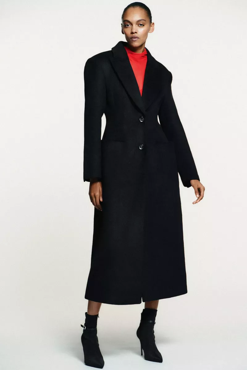 Woman in a long black wool coat and black high-heeled ankle boots stands in a studio against a white backdrop for a fashion editorial.