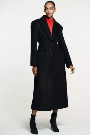 Woman in a long black wool coat and black high-heeled ankle boots stands in a studio against a white backdrop for a fashion editorial.