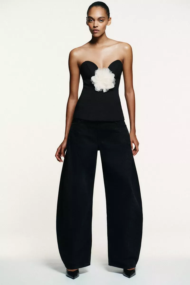 Elegant woman in a black strapless top with a white flower detail and wide-leg trousers posing in a studio setting.