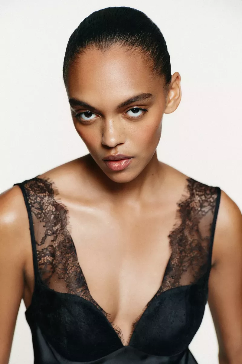 Close-up studio portrait of a woman with slicked-back dark hair wearing a black lace-trimmed top against a neutral backdrop.