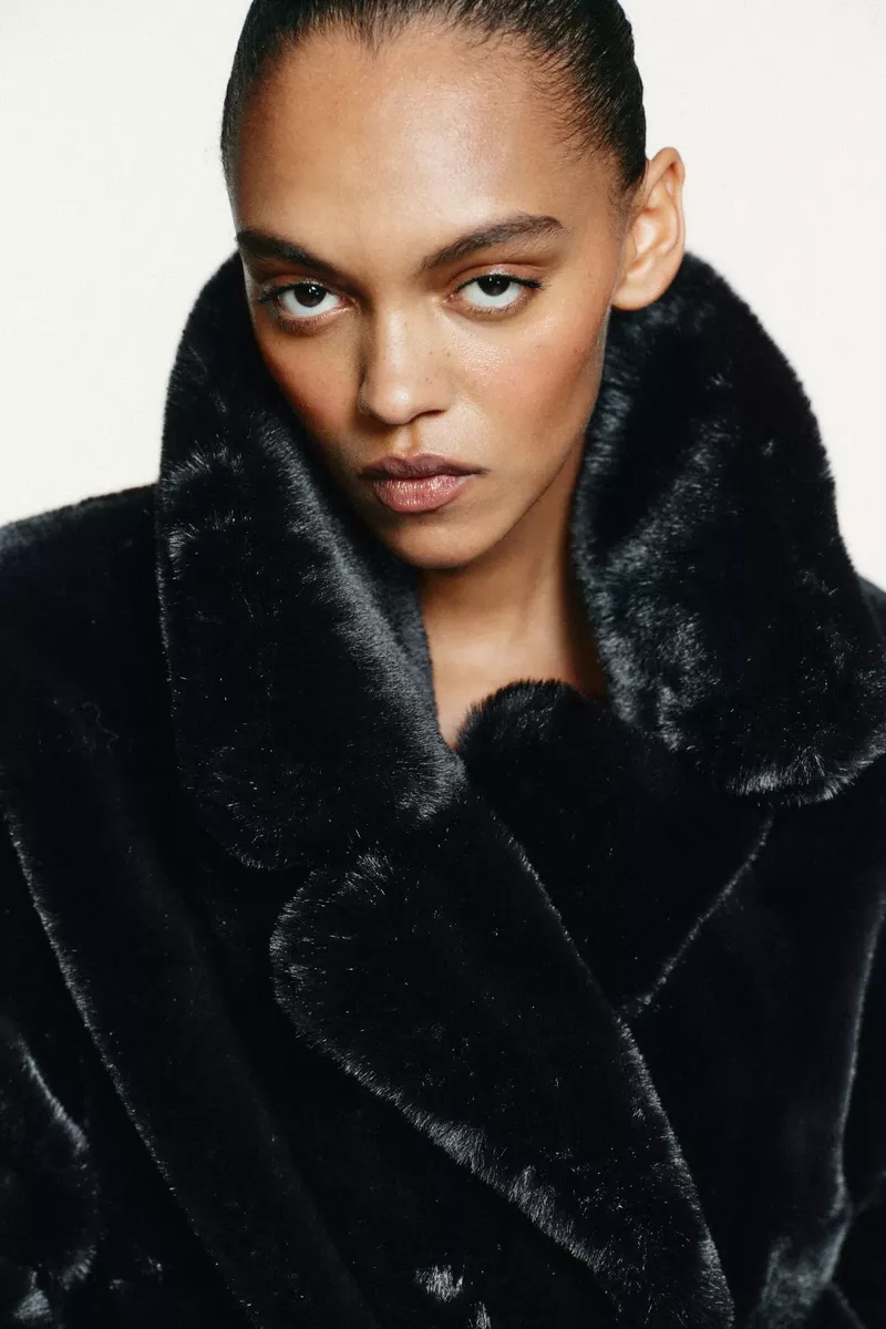 Close-up of a woman with fair skin wearing a black, faux fur coat. The portrait has a soft, diffused lighting with a white backdrop.