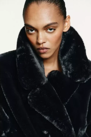 Close-up of a woman with fair skin wearing a black, faux fur coat. The portrait has a soft, diffused lighting with a white backdrop.