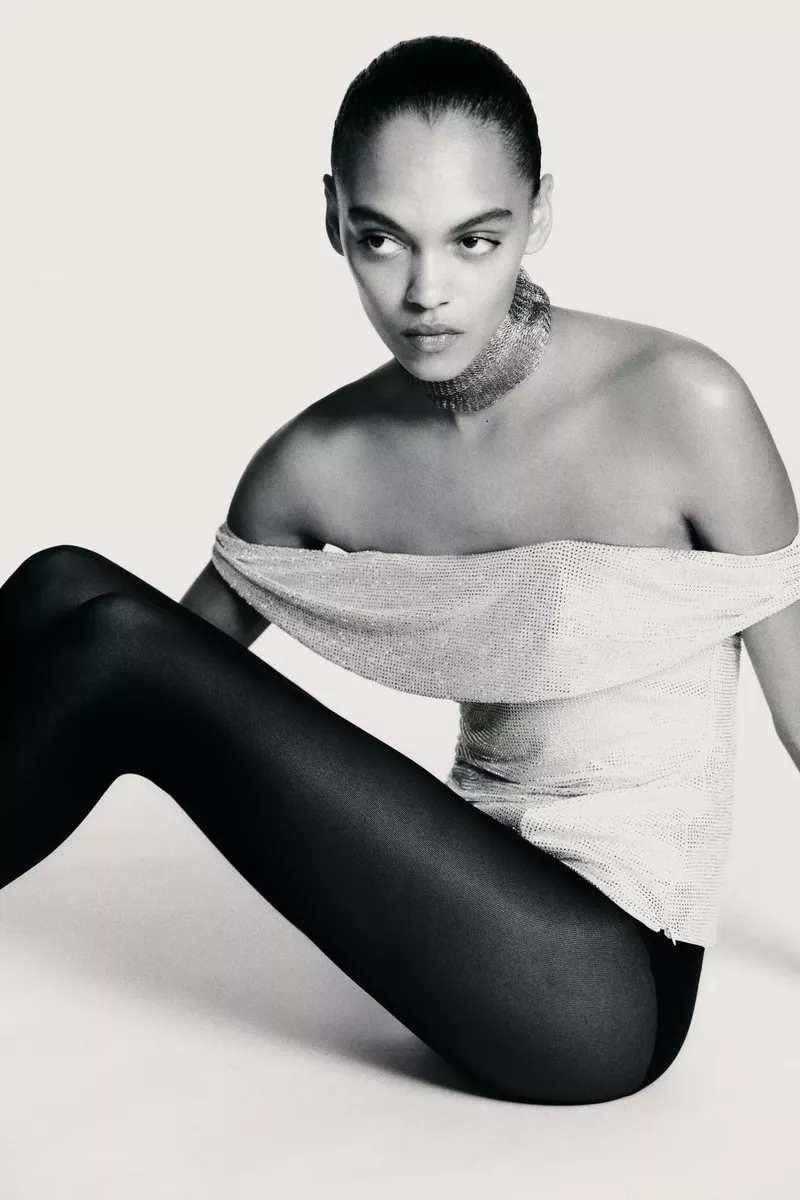 A model with an off-the-shoulder silver top and black tights sits in a studio against a white background in a fashion shot.