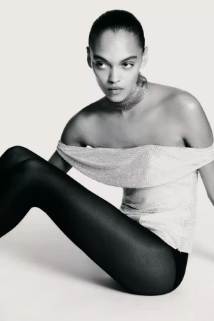 A model with an off-the-shoulder silver top and black tights sits in a studio against a white background in a fashion shot.
