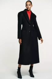 Model wearing a long black coat over a red top and black heeled boots against a white background for Zara.