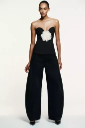 Model wearing a black strapless top with a white flower embellishment and wide leg trousers against a plain white background