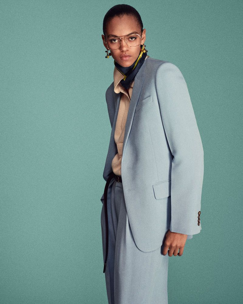 Professional portrait of a slender adult female wearing a blue suit, tan blouse, and stylish glasses against a solid teal studio backdrop.