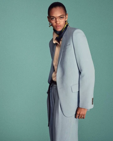 Professional portrait of a slender adult female wearing a blue suit, tan blouse, and stylish glasses against a solid teal studio backdrop.