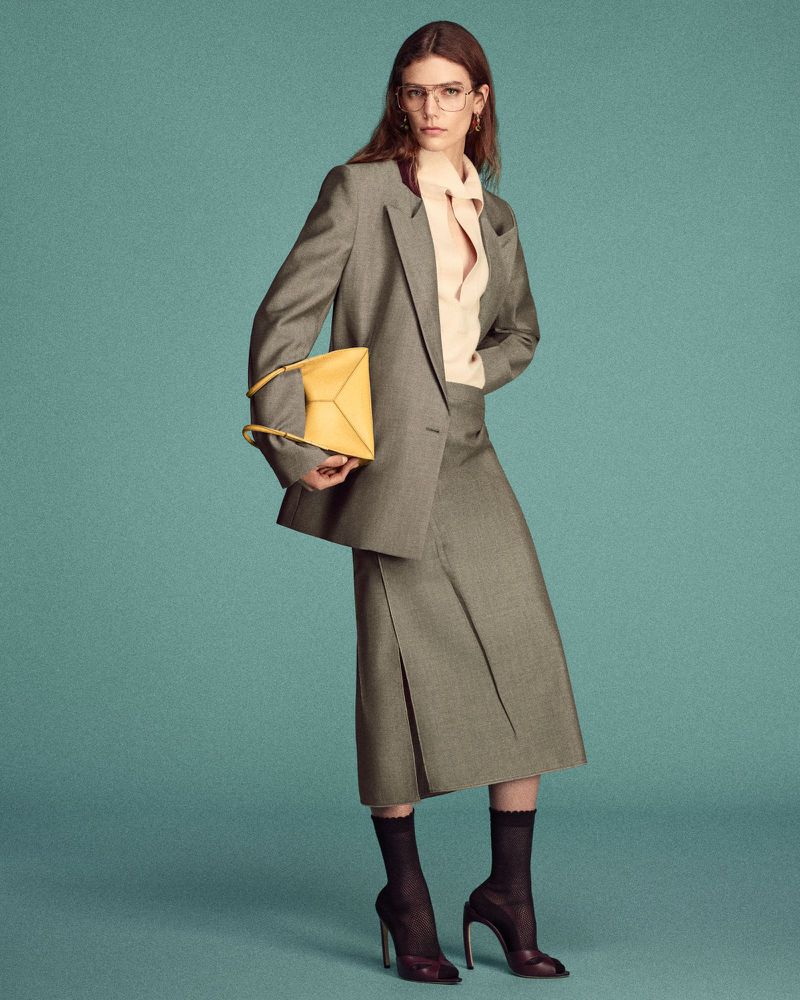 Woman in grey skirt suit poses with yellow handbag against teal backdrop. She wears black hosiery and glasses for a professional fashion look.