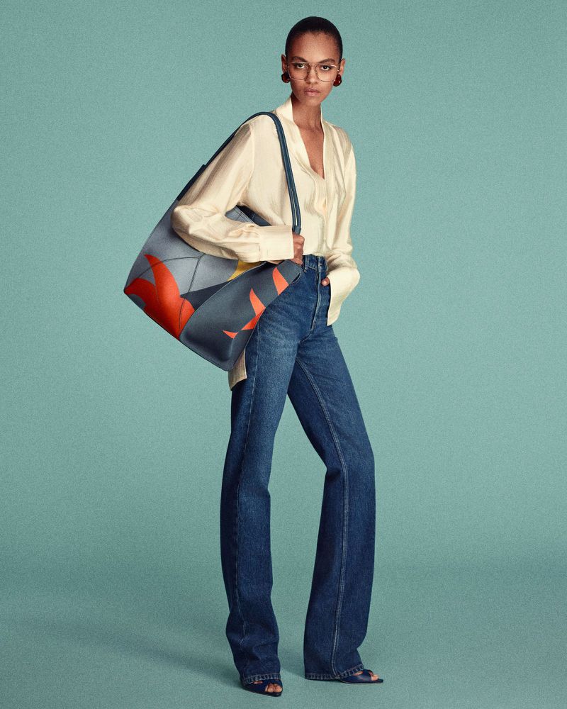 Fashion model poses in a studio with a patterned blue tote bag, wearing an off-white blouse, blue jeans, clear eyeglasses, and peep-toe shoes.