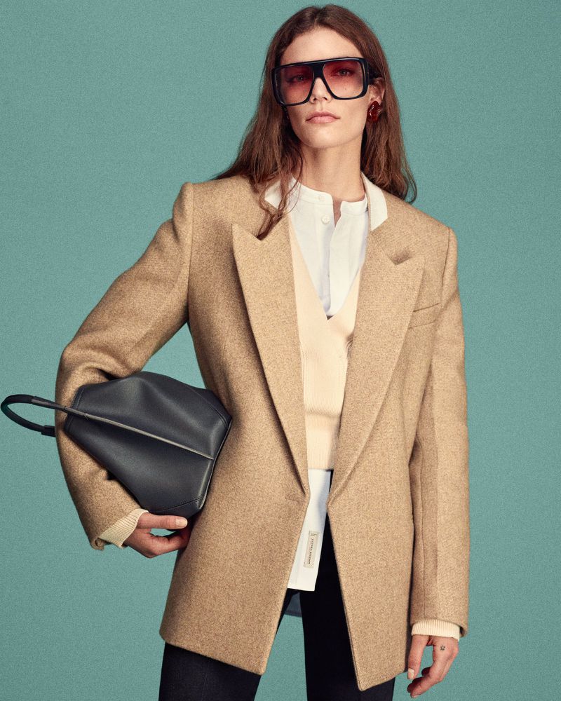 A fair-skinned woman with long hair poses wearing large sunglasses, a beige blazer, a white top, and holding a dark bag.