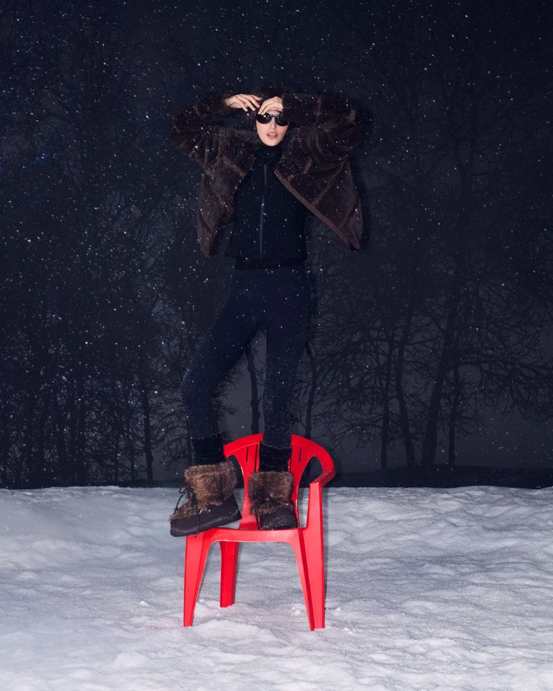 Fashionable woman in fur coat and sunglasses stands on a red plastic chair in a snowy field, against a dark starry background.