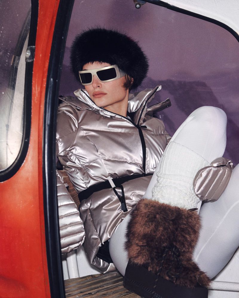 A woman in ski apparel and sunglasses sits inside a ski gondola with a snow-covered landscape visible in the background, fashion-focused.