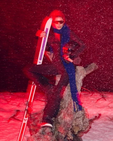 Woman in ski gear with skis poses on snowy tree stump against a red background in an outdoor, snowy setting with falling snow.
