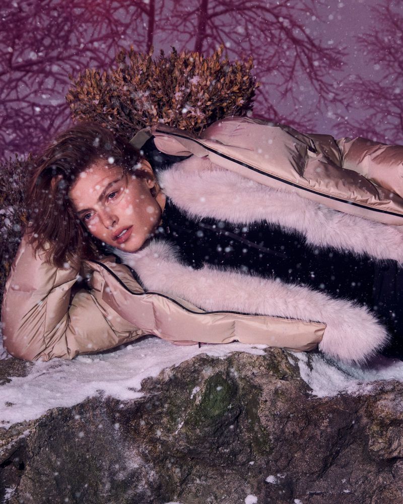 A woman in a warm coat poses outdoors in a snowy forest. She rests on rocky ground with trees and light snowfall in the winter season.